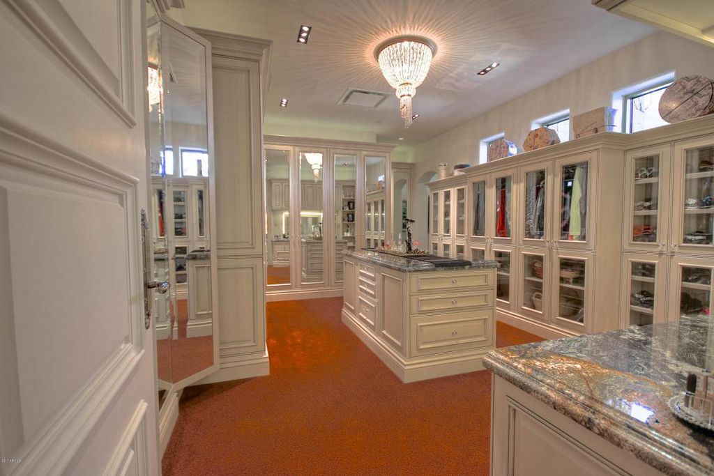 اتاق لباس بزرگ یک خانه لوکس که دارای ویترین های شیشه ای، کمد و کشوهای سفید و دیوارهای آینه کاری شده می باشد